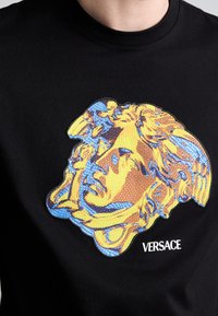 T-shirt preta com um gráfico texturizado e colorido de um rosto em tons de amarelo e azul. "Versace" está estampado em letras brancas abaixo.