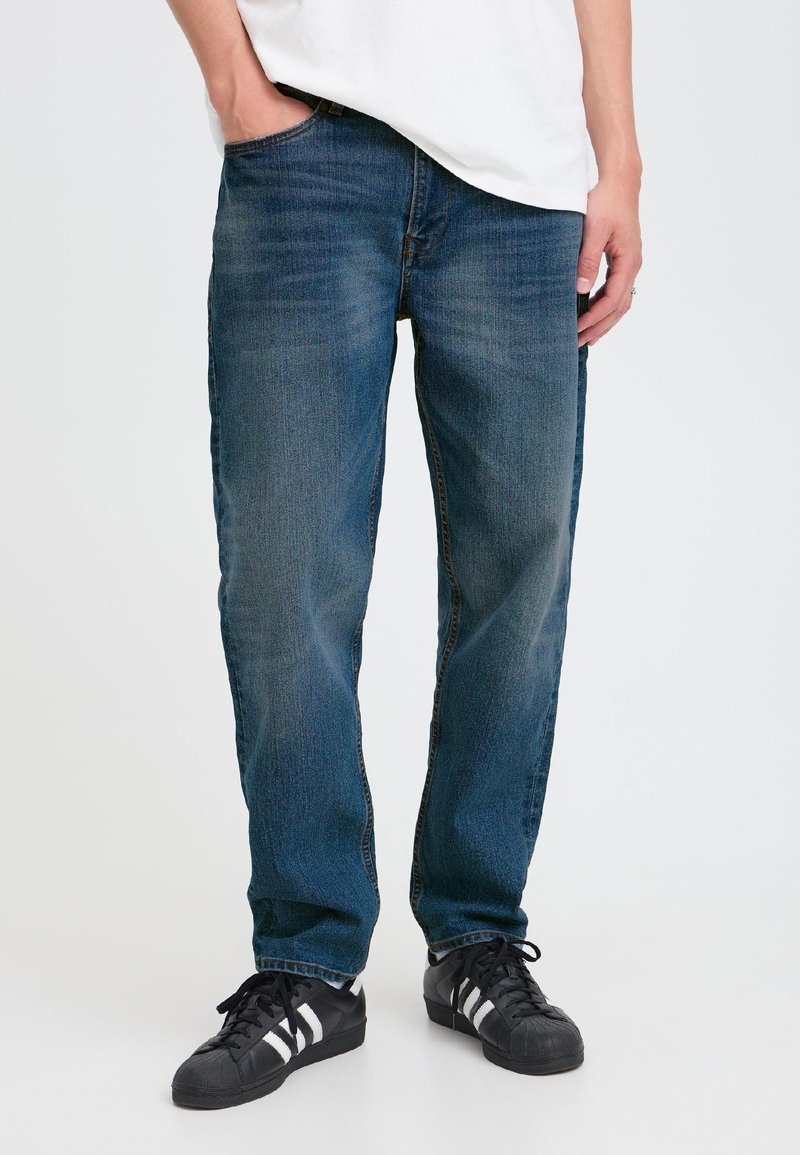 Persona che indossa jeans blu a gamba dritta e sneaker nere con strisce bianche, in piedi con una mano in tasca su sfondo bianco.