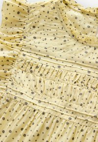 Żółta bawełniana sukienka z falbaniastym dekoltem i brązowym wzorem w kropki. Posiada pionowe zaszewki i marszczone sekcje wzdłuż gorsetu.