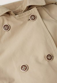 Beige trench coat-tyg med två bruna knappar, med en bred krage och rena sömmar. Slät textur utan synliga mönster.