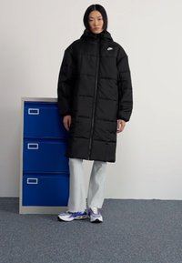 Melnā pūkaina jaka ar kapuci, ar priekšējo rāvējslēdzēju un izšūtu dizainu. Saskaņota ar gaismas krāsas plisētām biksēm un sporta apaviem.