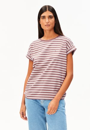 Vrouw met lang donker haar, gekleed in een mauve en witte gestreepte korte mouwen shirt en lichtblauwe spijkerbroek tegen een witte achtergrond.