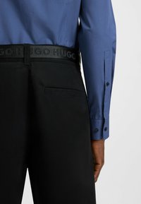 Pantaloni neri con una cintura testurizzata che presenta il logo "HUGO" ricamato. Camicia blu con polsini abbottonati, tessuto morbido e vestibilità sartoriale.