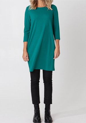 Femme portant une tunique sarcelle longueur genou avec manches trois-quarts, pantalon noir ajusté et bottines noires à lacets.