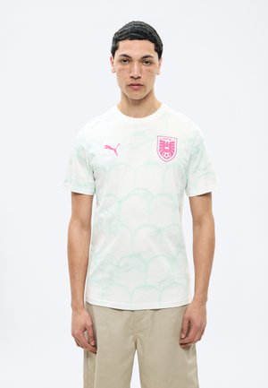 Bărbat tânăr cu păr scurt și creț, purtând un tricou alb de fotbal Puma cu logo roz și pantaloni bej, stând în fața unui fundal simplu.