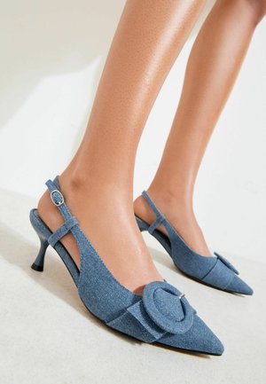 Fødder iført blå denim slingback kitten hæle med spidse tådele og store dekorative spænder på en lys gulv- og vægbaggrund.