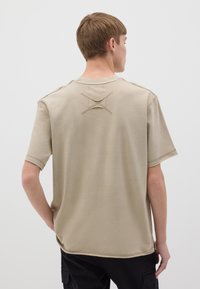 T-shirt beige dalla vestibilità ampia con maniche corte, caratterizzata da un dettaglio di punto croce sul retro. Tessuto in cotone morbido con un design semplice e pulito.