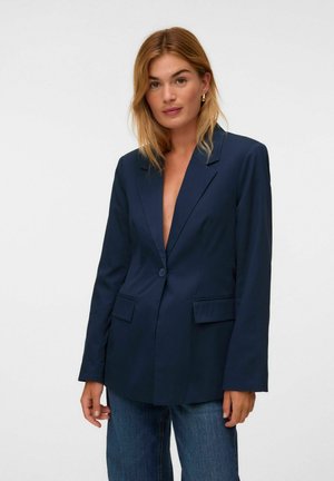 Femme aux cheveux blonds mi-longs portant un blazer bleu marine et un jean bleu, debout devant un fond clair uni.