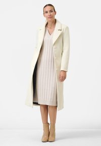 Long cream wool coat with notch lapels and two buttons over a beige ribbed knit dress. Model wears beige ankle boots.