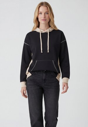 Woman with blonde hair wearing a black and beige hoodie and black jeans, standing against a plain light background.