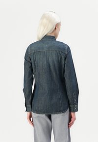 Tumšs džinsa krekls ar garām piedurknēm, pogu aprocēm un aizmugurē izšūtiem elementiem. Tam ir izliekts apakšmalas dizains un nedaudz pieguļoša forma.