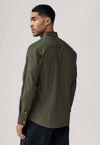 Olijfgroene overhemd met lange mouwen en een kraag. Glad textuur, met knoopmanchetten en een lichte glans. Bevat een middennaad aan de achterkant.