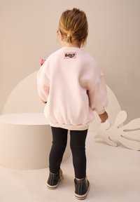 Sweat-shirt oversize rose clair avec poignets et ourlet côtelés, orné d'un logo "Bluey" au dos, accompagné de leggings noirs et de chaussures noires.