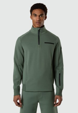 Man wearing olive green half-zip pullover with zippered chest pocket and matching pants, standing against plain gray background.