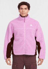 Häggig fleecejacka med en ljusrosa kropp och mörkbruna ärmar, ståkrage, hel dragkedja och logotyp på bröstet.