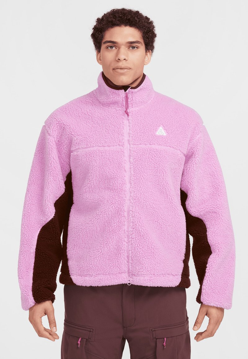 Häggig fleecejacka med en ljusrosa kropp och mörkbruna ärmar, ståkrage, hel dragkedja och logotyp på bröstet.