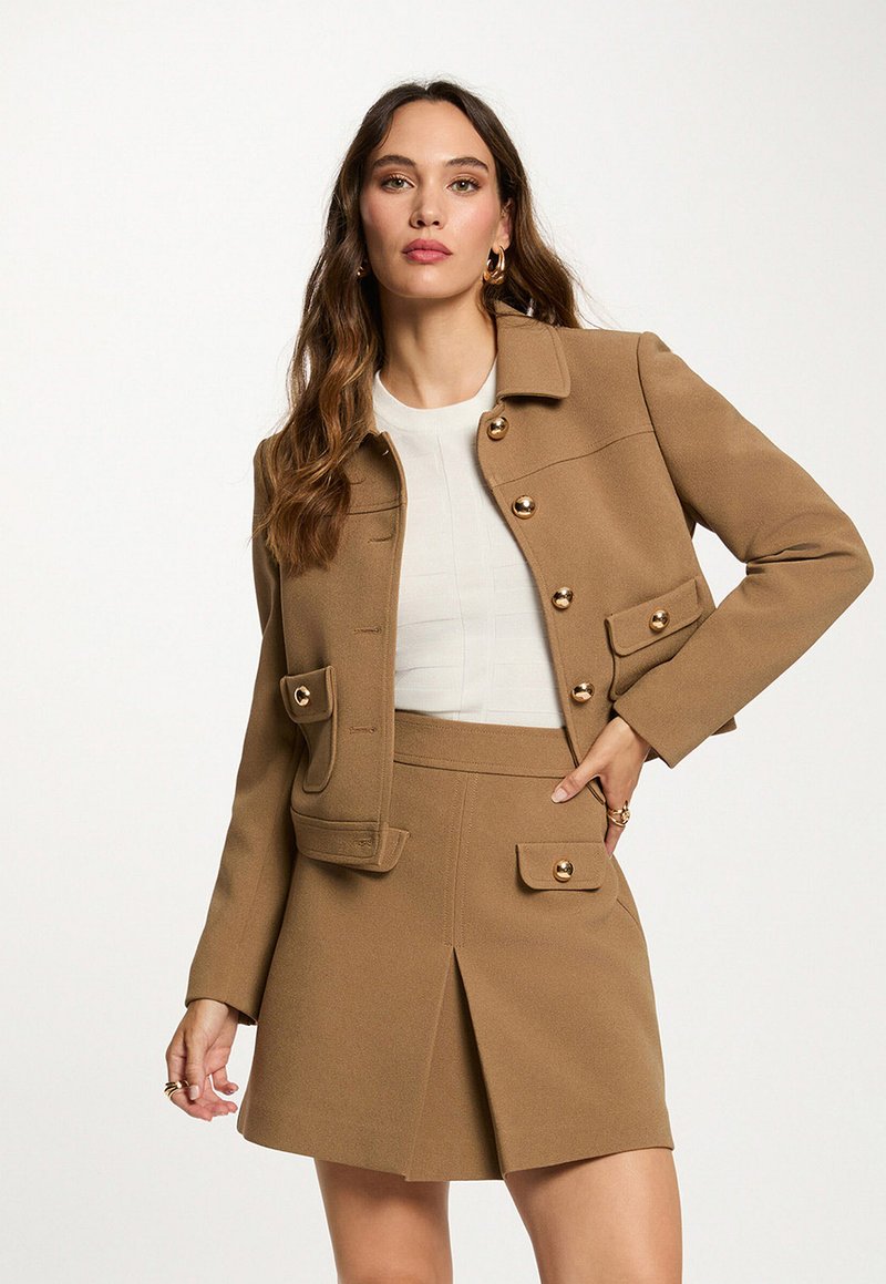 Vrouw in een camel-kleurige, knoop-jack en bijpassende rok met gouden knopen, gecombineerd met een witte top, poseert tegen een witte achtergrond.