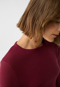 Bordeaux geribbelde top met ronde hals, met een gestructureerd oppervlak en een getailleerde silhouet, getoond van de schouders omhoog, met aandacht voor het haar.