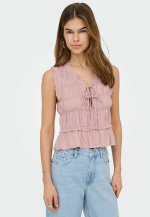 Fiatal nő hosszú barna hajjal, rózsaszín ujjatlan, fodros felsőt és világoskék farmert visel, egyszínű háttér előtt áll.