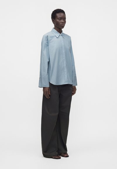 Femme portant une chemise bleu clair oversize à manches longues, un pantalon noir à jambes larges et des sandales noires à bouts ouverts, debout sur un fond uni.