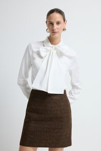 Witte blouse met een grote strik bij de kraag, gecombineerd met een bruine tweed minirok met een gestructureerd patroon en metallic accenten.