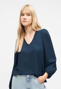 Νεαρή γυναίκα με μακριά ξανθά μαλλιά, ντυμένη με μπλε μπλούζα V-λαιμόκοψη με υφή και ανοιχτόχρωμο μπλε τζιν, με το ένα χέρι στην τσέπη.