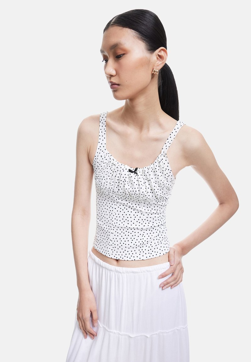 Hvid tanktop med prikker, der har en samling i sømmen og en sort sløjfe ved halsudskæringen, parret med en svævende hvid nederdel.