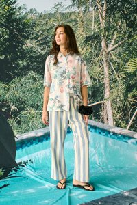 Femme debout devant un décor de piscine portant une chemise fleurie à manches courtes, un pantalon large rayé, un chouchou noir et des tongs noires.