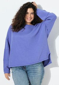 Mujer sonriente con cabello rizado lleva un suéter morado suelto y jeans azul claro, posando con una mano en el cabello contra un fondo blanco.
