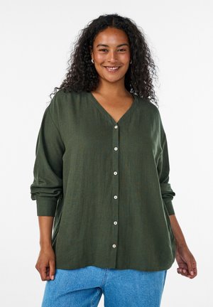 Femme aux cheveux bouclés souriante, portant un chemisier boutonné vert olive et un jean bleu clair, debout devant un fond blanc.
