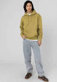 Jeune homme aux cheveux bouclés portant un sweat à capuche vert olive, un jean gris clair et des chaussures marron, debout devant un fond gris clair uni.