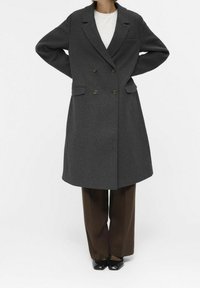 Abrigo gris de lana de corte doble con solapas acanaladas, dos bolsillos frontales y botones de latón; combinado con pantalones marrones y zapatos negros.
