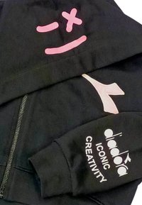 Felpa nera con zip caratterizzata da design rosa e crema: un volto sorridente, un logo stilizzato e il testo "diadora ICONIC CREATIVITY" sul maniche.