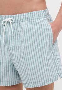 Hellblaue Badehose mit vertikalen Streifen, elastischem Bund und Zugband. Sie verfügt über Seitentaschen und ein strukturiertes Material.