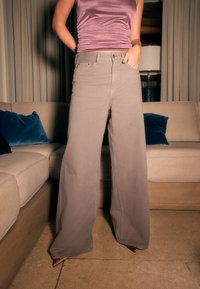 Jasnoszare dżinsy o szerokich nogawkach z wysokim stanem i tradycyjnym krojem z pięcioma kieszeniami, zestawione z dopasowanym różowym topem, na tle neutralnego wnętrza.