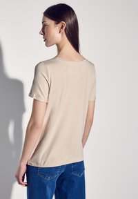 T-shirt beige à manches courtes avec un col rond, texture lisse et ourlet légèrement froncé, associé à un jean en denim bleu.