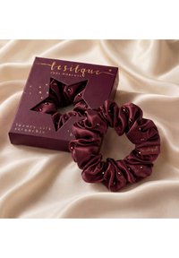 Donkerrood zijden scrunchie met kleine kralen, naast een bijpassende merkdoos met stervormig venster, geplaatst op lichte satijnen stof.