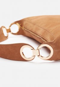 Anse de sac à main en daim marron avec deux grandes boucles dorées entrelacées et un détail de fermeture éclair sur un sac en daim assorti.