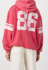 Sudadera roja de tela con un aspecto desgastado, que presenta un gran "86" en la parte posterior y rayas blancas en las mangas. Diseño acogedor y de corte oversize.