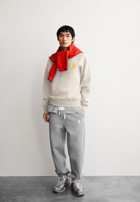 Jonge man in een beige sweatshirt en grijze joggingbroek met een rood sweatshirt over zijn schouders, staand tegen een eenvoudige witte muur.