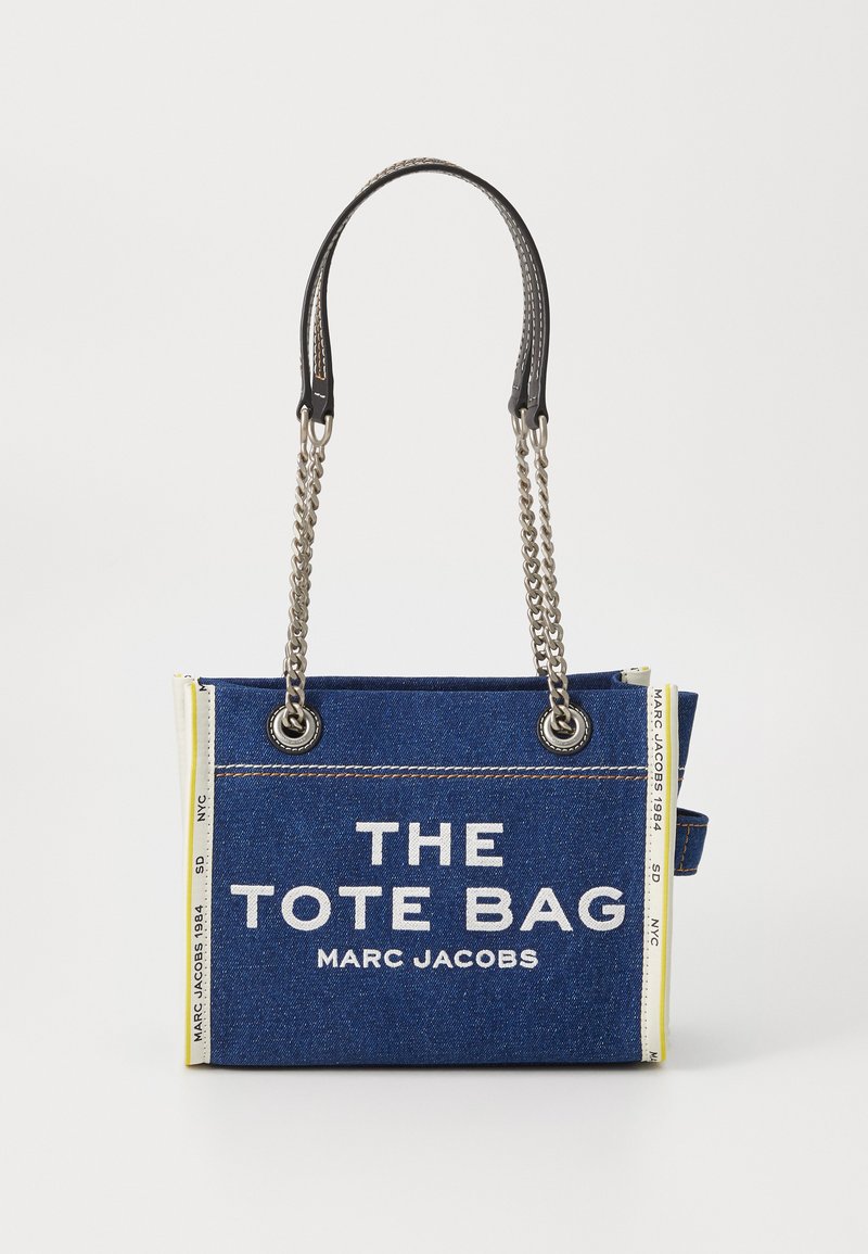Džinsu iepirkumu soma ar melnu ādu un sudraba ķēdes siksnām. Iekļauj baltu tekstu ar uzrakstu "IEPIRKUMU SOMA MARC JACOBS" uz zila fona.