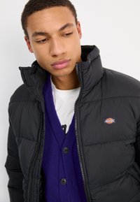 Giubbotto imbottito nero con colletto alto, caratterizzato da una patch con logo. Indossato sopra un cardigan viola con bottoni, sovrapposto a una camicia bianca.