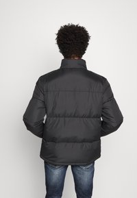 Giubbotto imbottito nero con colletto alto, caratterizzato da sezioni trapuntate e una trama liscia e lucida. Indossato sopra dei jeans su uno sfondo neutro.