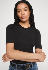 Black short-sleeve T-shirt with a round neckline. The fabric is smooth and fitted, paired with blue denim pants and a black belt with a gold buckle.