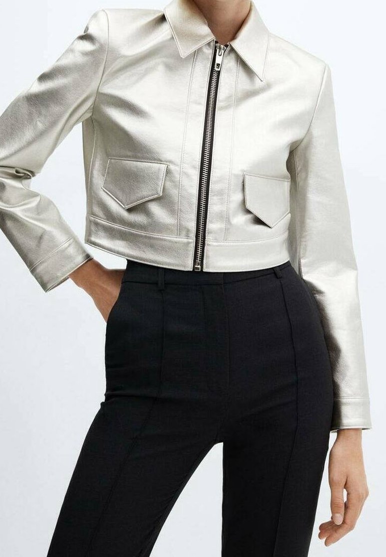 Veste argentée brillante coupée avec fermeture éclair devant et poches à rabat, portée avec un pantalon noir taille haute, une main dans la poche.