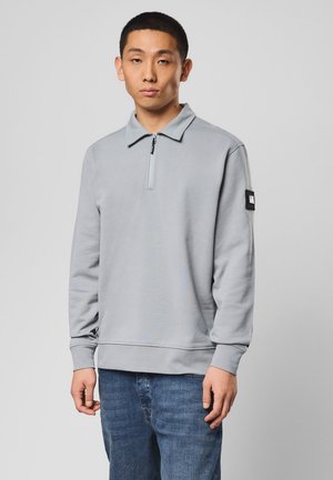 Młody mężczyzna ubrany w jasnoszary pulower z zamkiem do połowy długości i czarną łatą na lewym rękawie oraz niebieskie jeansy, stojący na jednolitym tle.