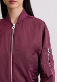 Woman wearing a dark purple bomber jacket with front zipper and zippered pocket on the upper sleeve, against a plain light wall.