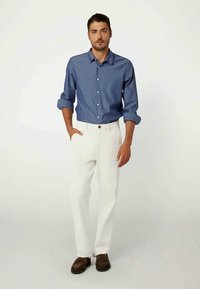 Camisa azul de botões, mangas enroladas, calças brancas, mocassins castanhos. O conjunto apresenta um corte clássico com uma aparência limpa e casual.