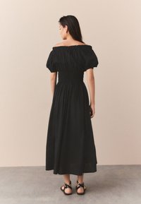 Schwarzes schulterfreies Kleid mit Puffärmeln, geraffter Taille und knöchellangem Rock. Kombiniert mit schwarzen Riemchensandalen.