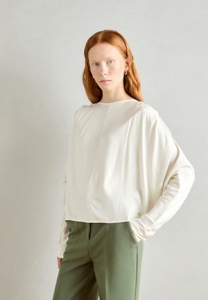 Lässig geschnittene, elfenbeinfarbene Bluse mit rundem Halsausschnitt und langen Ärmeln, kombiniert mit olivgrünen Hosen. Softe Textur, minimale Details.